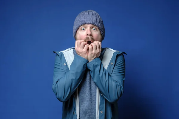 Überraschter, schockierter Mann mit Strickmütze und wasserdichter Jacke ist verängstigt, öffnet erstaunt den Mund und blickt mit großen Augen. Vereinzelt auf blauem Hintergrund. — Stockbild Schockierter Mann mit Strickmütze und wasserdichter Jacke ist verängstigt, öffnet erstaunt den Mund und blickt mit großen Augen. — Stockfoto