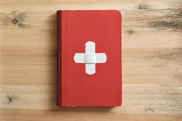 Old red book with a white cross looks like the flag of Switzerland, on a wooden table background. Creative idea. Top view.