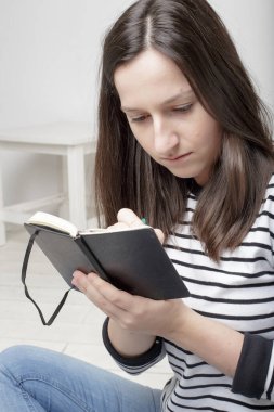 Not defterine kalem kullanarak notlar alan, beyaz ahşap zeminde oturan genç ve güzel bir kız öğrenci.