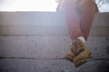 Şık giyimli bir kız, bahar güneşinin ılık ışınlarında beton bir çitin üzerinde sallanarak ve bacak bacak bacak üstüne atarak oturur..