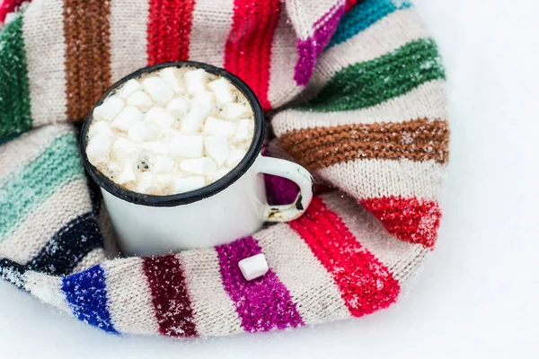 Old metal cup of hot cocoa with marshmallows and a soft colorful knitted scarf on a fluffy the snow close-up