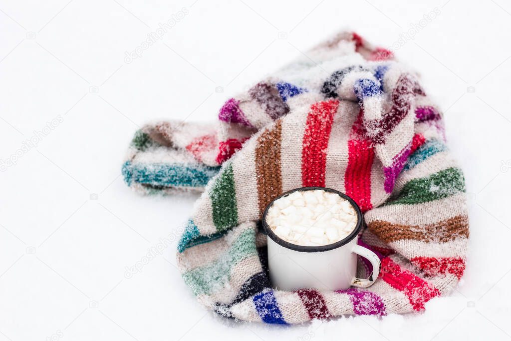 Old metal cup of hot cocoa with marshmallows and a soft colorful knitted scarf on a fluffy the snow