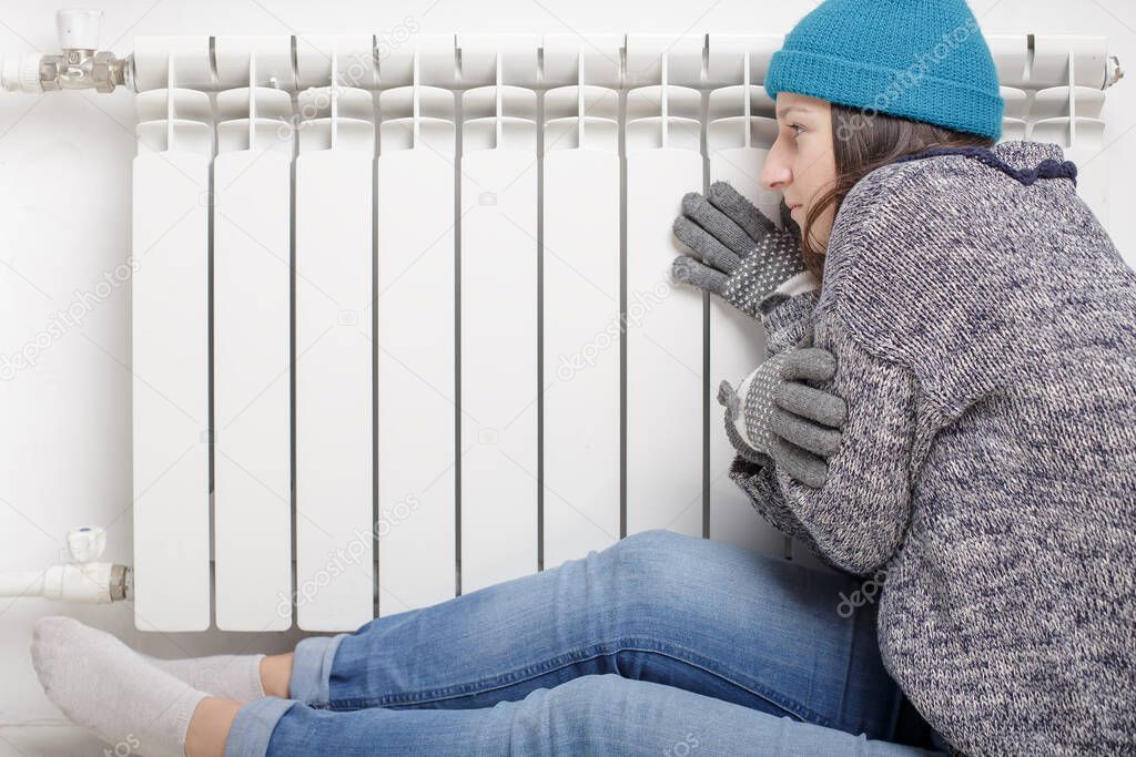 Young beautiful girl froze and warms hands near a radiator, dressed in gloves, a sweater and a hat