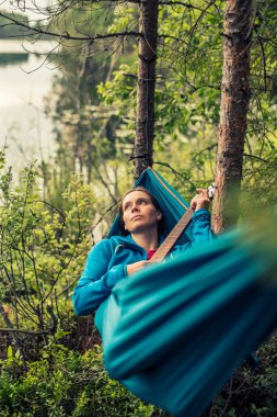 Düşünceli güzel kız bir hamakta uzanıyor ve gitar melodisini tıngırdatıyor, bir yaz akşamı ormanda gölün arka planında.