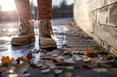 Botlu bacaklar, güneş ışınları altında, buzla kaplı bir su birikintisinde kayadan yapılmış kaldırımın üzerinde, düşmüş yapraklar üzerinde duruyor. Yakın plan..