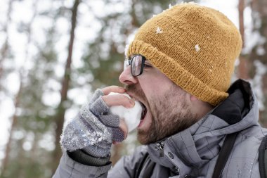 Genç sakallı bir adam ormanda zevkle kartopu yer..