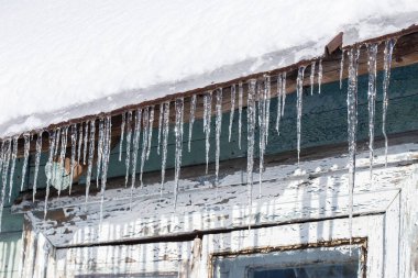 Eski bir evin çatısında asılı duran buz saçakları. Kırsal kesimde güneşli bir bahar günü boyaları dökülüyor..