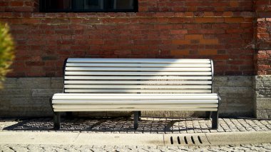 Ilık, güneşli bir bahar gününde, eski bir tuğla binanın zeminine taş bir kaldırıma düşen boş beyaz bir bank..