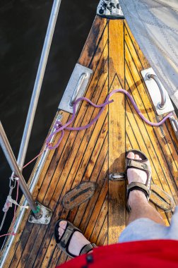 Karanlık suyun arka planındaki bir yatın ahşap güvertesinde duran sandaletli adam. Yakın plan..