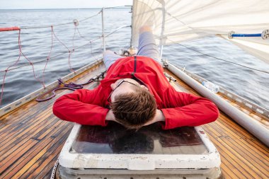 Kırmızı ceketli ve gözlüklü bir adam hareket halindeki bir yatın güvertesinde, kollarını başının altına katlıyor, denizin ve gökyüzünün arka planına karşı, bulutlu..