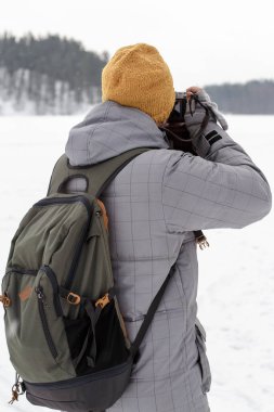 Sırtında sırt çantası ve kalın giysiler içinde elinde kamera olan bir adam kış manzarasının fotoğraflarını çekiyor. Karla kaplı kayaların, ormanların ve donmuş göllerin arkaplanına karşı, kış günü bulutlu..