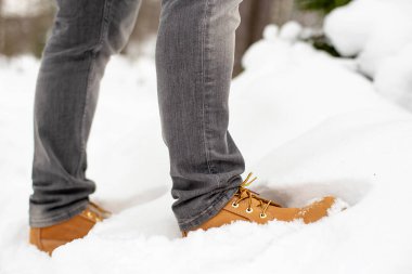 Modaya uygun sarı ayakkabılı bir adamın bacakları kar yığınında duruyor, karla kaplı bir ormanın bulanık arka planına karşı. Yakın plan..