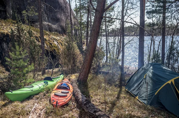 Two kayaks are in the spring forest near the camp, the sun and the smoke from the fire, near the lake and tents.