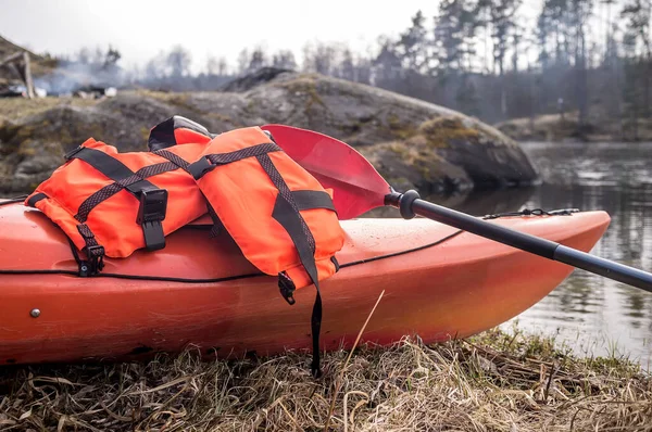 An orange life jacket and paddle lie on a kayak that stands on a borega of a lake, against a background of fire and rocks. Close-up.