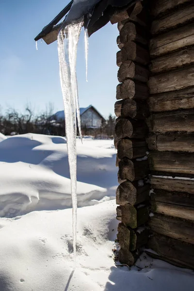 強力で長い透明なつららは 古いログハウスの屋根の端からぶら下がり 雪のドリフトに達します 太陽の裏側では清々しい春の日 — ストック写真