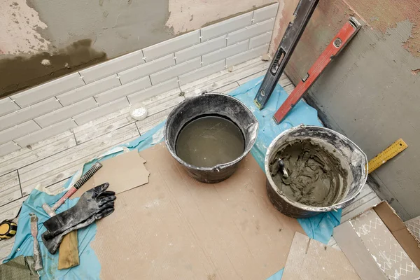 Repair in the bathroom. Several rows of tiles are laid on the wall with the help of a solution that is in the bucket and the tool.