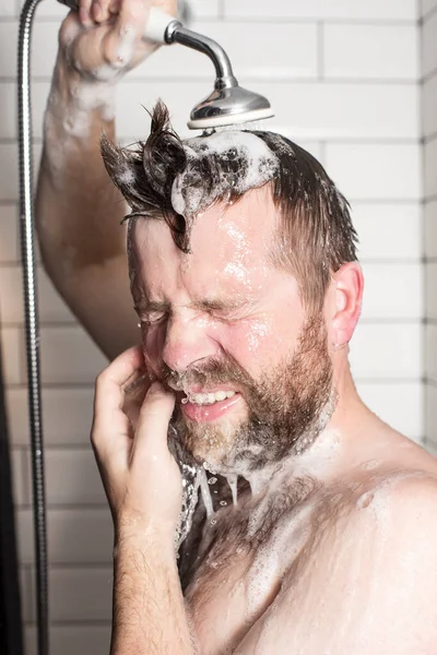 A handsome, bearded man washes the shampoo from his hair in the bathroom with a shower head with warm running water.