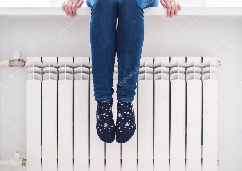 The girl sits on the windowsill and warms her feet in pretty socks at the radiator, on a cold winter day.