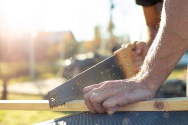Bir adam, bir marangoz el testeresi ile ahşap bir ürün kesiyor. Sıcak bir yaz gününde, açık havada..