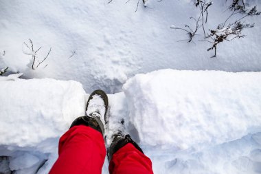 Sıcak botlar içindeki insan bacakları, güneşli bir kış gününde, küçük bir derenin üzerinden geçen alçak bir köprüde kar altında duruyor..
