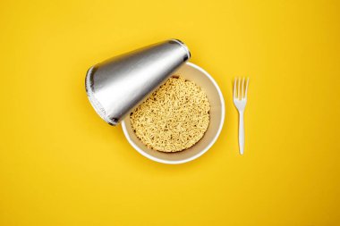Susuz bir fincanda nefis hazır erişteler ve parlak sarı bir arka fona yerleştirilmiş açık bir kapakla plastik bir çatal var. Üst görünüm.