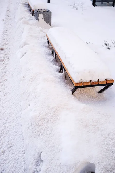 metal bench with a wooden seat covered with snow in a park near the footpath