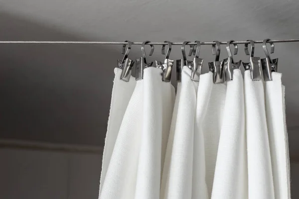 A cream curtain hangs on a rope on a metal hook, against the background of the ceiling and wall, illuminated by daylight.