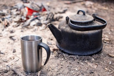 Sıcak bir bardak ve yerde bir çaydanlık. Ateşin yanında, güneşli ve sıcak bir kampta..