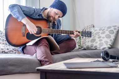 Odaklanmış genç besteci, akustik bir gitarla, bir deftere bakarken ve ağzında bir kalemle, rahat bir ev ortamında, rahat bir kanepede otururken,.