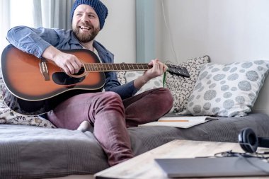 Genç besteci, notaları akustik gitarla çalıyor, sonuçtan memnun, arkadaşlarına mutlu bir şekilde gülümsüyor, pencereden gelen doğal ışıkla rahat bir ev ortamında..