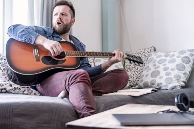 Genç bir erkek akustik gitar çalar ve kendi bestesini söyler. Bir kanepede, rahat bir ev ortamında, pencereden doğal bir ışık altında oturur.