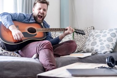 Genç, yakışıklı bir erkek besteci akustik gitar çalar ve yüksek sesle kendi bestesini söyler, bir kanepede oturur, rahat bir ev ortamında, pencereden gelen doğal ışıkla.