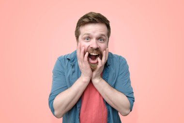 A happy, astonished Caucasian man with a beard, emotionally holds his hands on his face and opened his mouth from a pleasant surprise. Isolated on coral background. Close-up. clipart