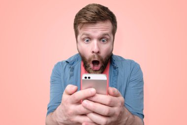 Şaşırmış, şok olmuş sakallı bir adam elinde bir akıllı telefon tutuyor ve ağzı açık ve büyük gözleriyle ekrana bakıyor. Mercan zemininde izole edilmiş. Moda renkleri. Yakın plan..