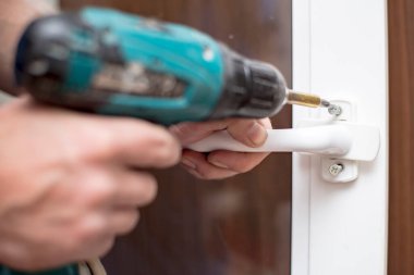 Adam elinde bir elektrikli tornavida tutuyor ve PVC penceresinin sapını vidalıyor. Yakın plan..