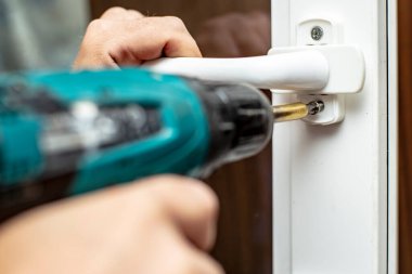 Adam elinde bir elektrikli tornavida tutuyor ve PVC penceresinin sapını vidalıyor. Yakın plan.