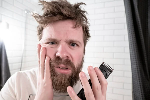 A man holds a trimmer in his hands looking at his reflection in the mirror and is horrified by his shaggy hairstyle and beard, early in the morning, against the background of a wall in the bathroom.