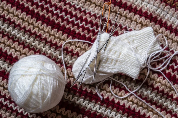 A clew of white wool yarn, knitting needles and unfinished mittens lie on a homemade multi-colored bedspread. Close-up