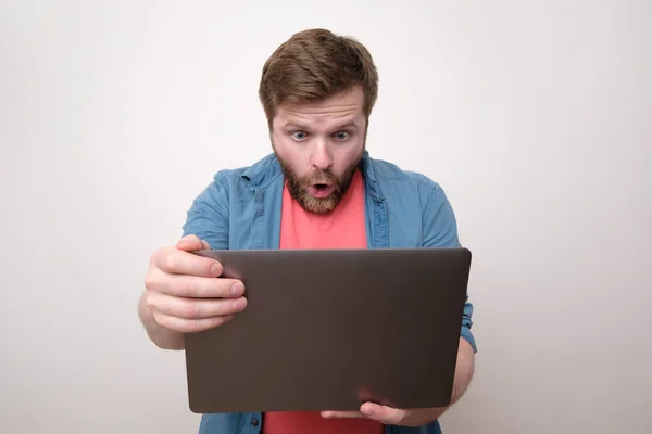 A shocked bearded man holds a laptop in his hands and looks at him with surprise with his mouth open and big eyes. Isolated on white background. Close-up.