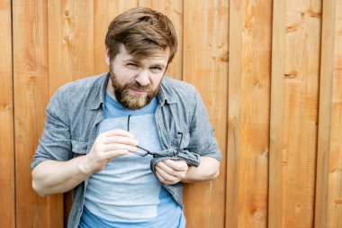 Serious bearded man cleans glasses by rubbing them with the help of his shirt and squinting, looking at the camera with hostility. Isolated on the background of an old wooden wall. Close-up. clipart