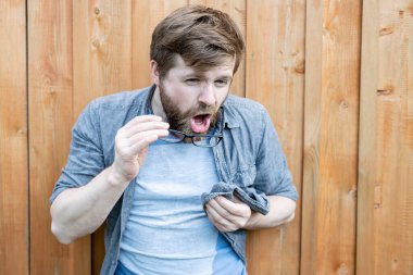 Bearded man cleans his glasses from dust and is going to wipe them with a shirt. Isolated on the background of an old wooden wall. Close-up. clipart