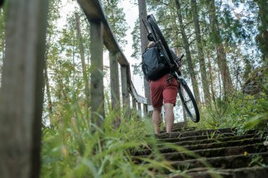 Sırt çantalı güçlü bir adam bisikletini omzunda tutar ve merdivenleri tırmanır, çimlerle kaplı, çam ağacının arka planına karşı..