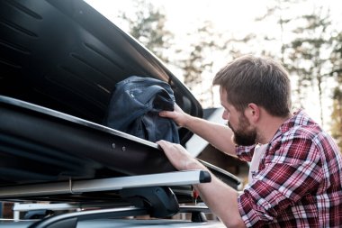 Bir adam eşyaları bir arabanın tavan rafına ya da bir kargo kutusuna koyar. Bir yaz akşamında evin ve ağaçların bulanık arka planına. Ortak aile gezisi. Yakın plan..