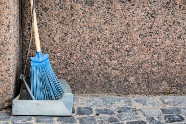 Metal kepçe ve tahta saplı plastik süpürge granit duvarın yanındaki kaldırımda duruyor..