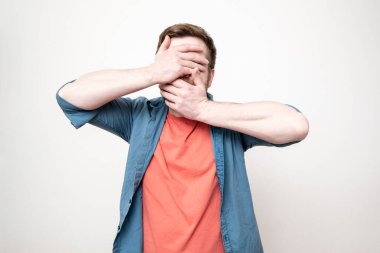 Kırmızı tişörtlü ve mavi gömlekli bir adam elleriyle yüzünü kapatıyor ve dirseklerini farklı yönlere açıyor. Beyaz arkaplanda izole.