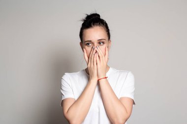 Şok olmuş, üzgün genç bir kadın iki eliyle ağzını kapattı ve yüzünü çevirdi. Beyaz bir arkaplanda izole edilmiş.