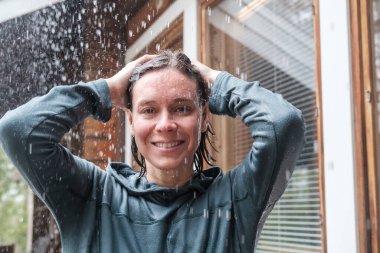 Sıcak yağmurun tadını çıkaran mutlu bir kadın. Geniş bir gülümsemeyle evin arka planında kameraya bakıyor..