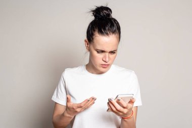 Şok olmuş, kafası karışmış genç bir kadın akıllı telefonuna bakar, kötü haberleri ya da fotoğrafları görür, kaşlarını çatar ve elle karışık bir jest yapar. Olumsuz duygular. Beyaz arkaplanda izole.