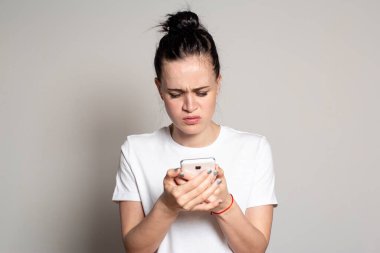 Üzgün genç bir kadın akıllı telefonuna bakar ve kaşlarını çatar. Olumsuz duygulara neden olan kötü haberler ya da fotoğraflar görüyor. Beyaz arkaplanda izole.