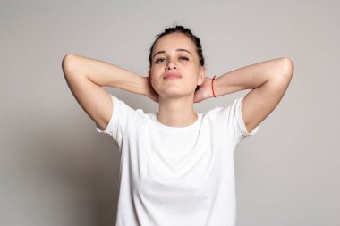 Güzel, sakin genç bir kadın elleri başının arkasında katlanmış ve sabit, kendinden emin bir bakışla kameraya bakıyor. Beyaz arkaplanda izole.